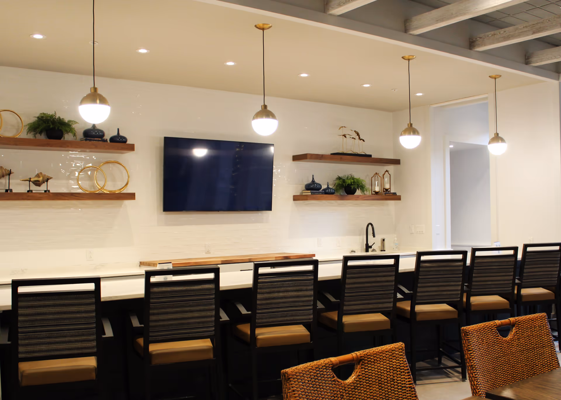 Modern communal kitchen/bar area with a long counter, a row of bar stools, pendant lights, a wall-mounted TV, and decorative shelves.