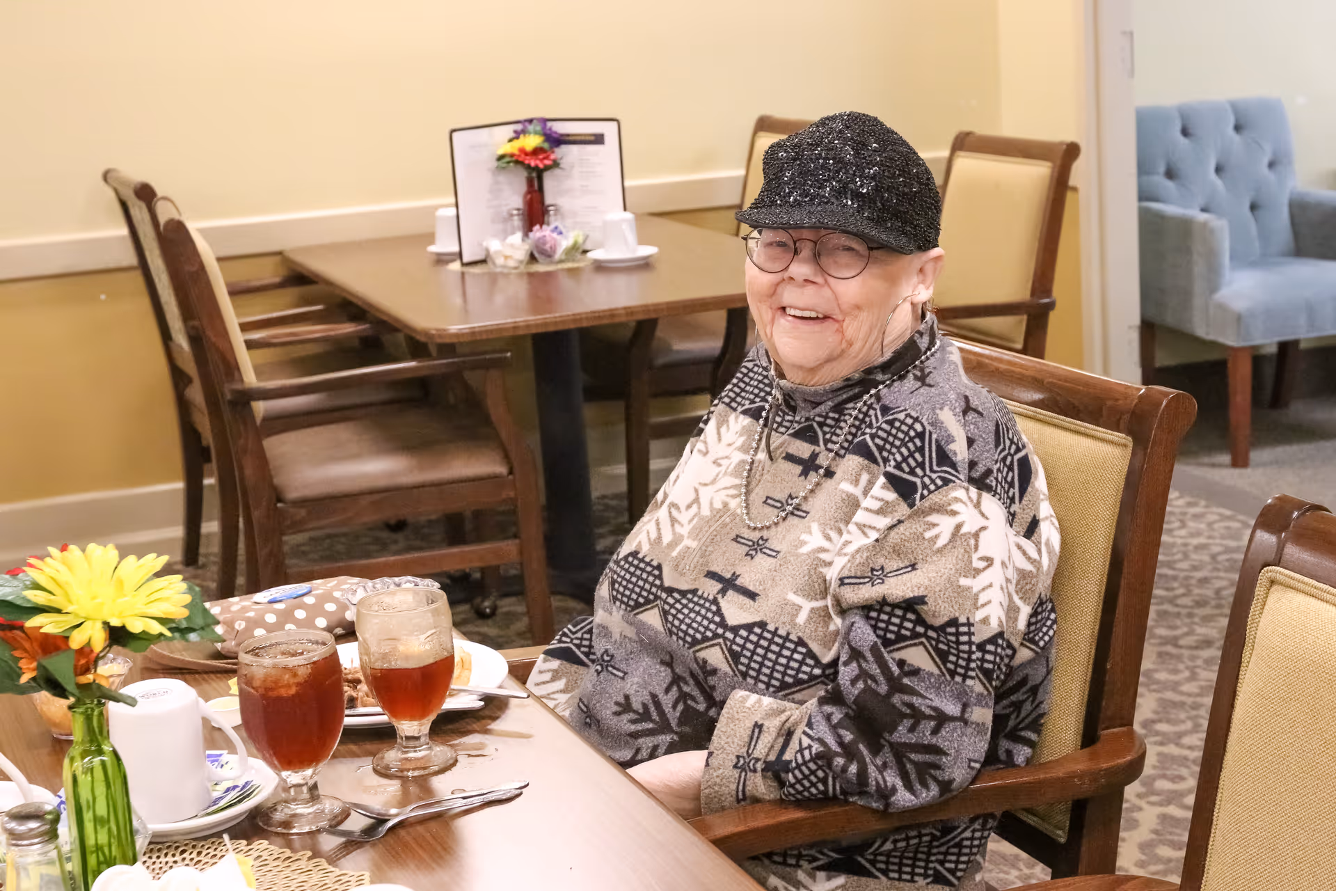 An elderly woman wearing glasses, a black sequined cap, and a patterned sweater is sitting at a dining table with two glasses of iced tea and a plate of food. The dining area has wooden chairs and tables, with a small flower arrangement on the table.