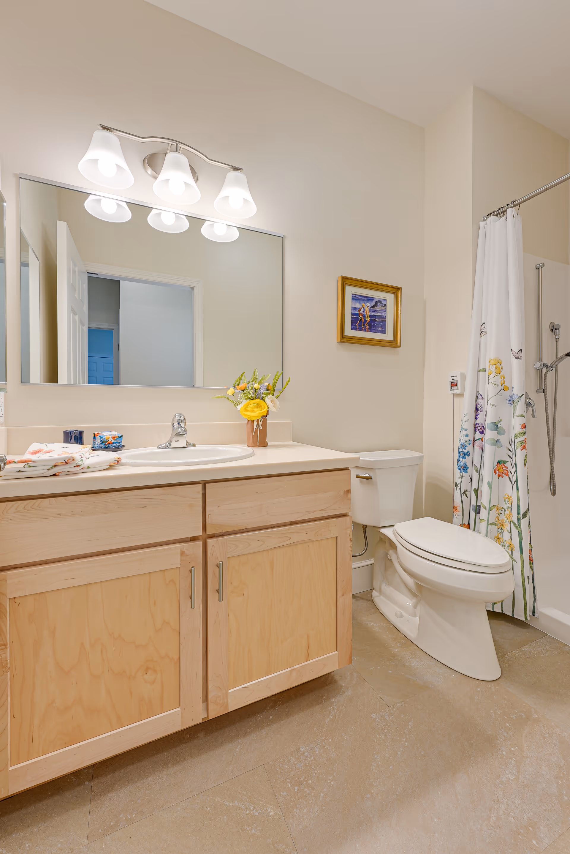 Well-lit bathroom with a wood vanity and sink, large mirror, toilet, and a floral shower curtain.