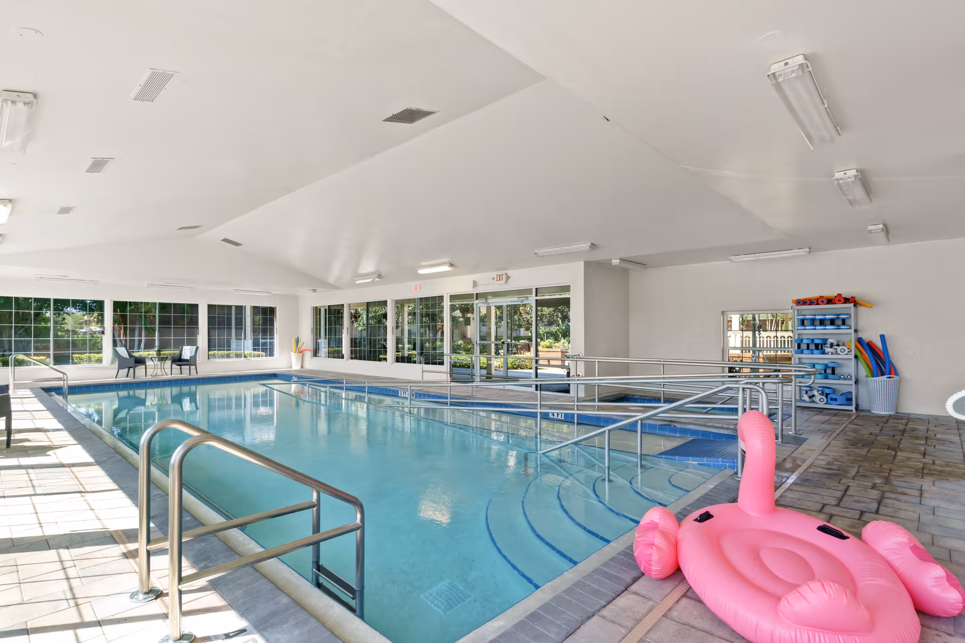 Indoor swimming pool area with handrails and steps leading into the water. There is a pink inflatable flamingo float on the tiled floor near the pool. Large windows and glass doors allow natural light to fill the space, and pool equipment is stored on shelves in the corner.
