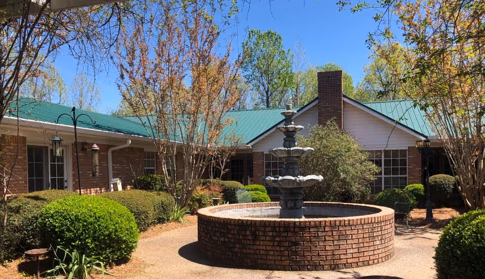 Outdoor courtyard area of a facility with a circular brick fountain in the center, surrounded by trimmed bushes and trees. The building has a green metal roof and brick walls with windows and a chimney visible. The sky is clear and blue.