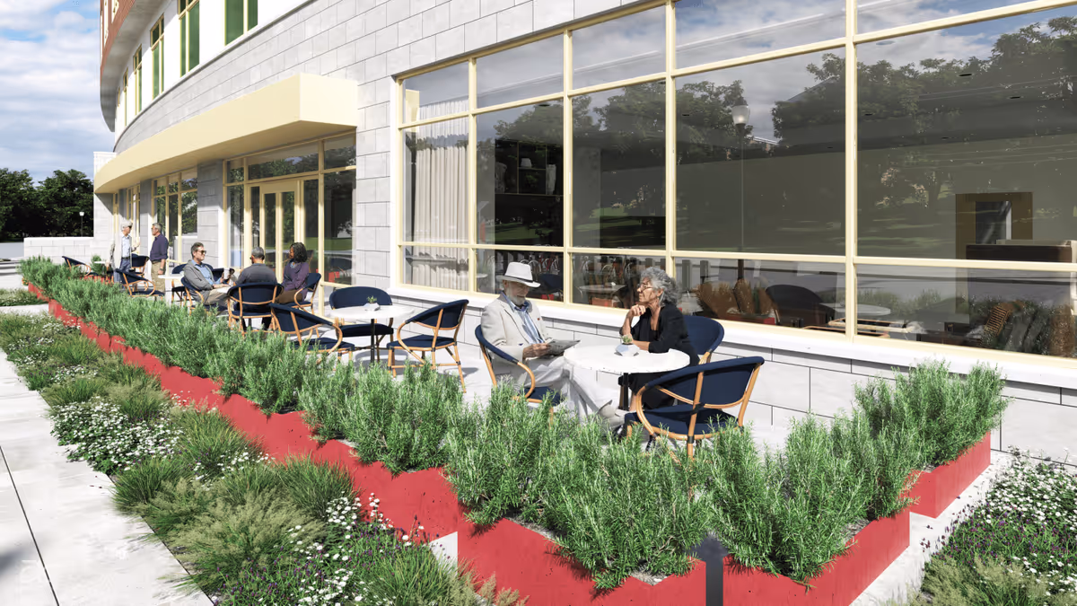 Outdoor patio area at Benchmark at Alexandria with several elderly people sitting at round tables with chairs, surrounded by green plants and red planters, adjacent to a building with large windows reflecting trees and sky.