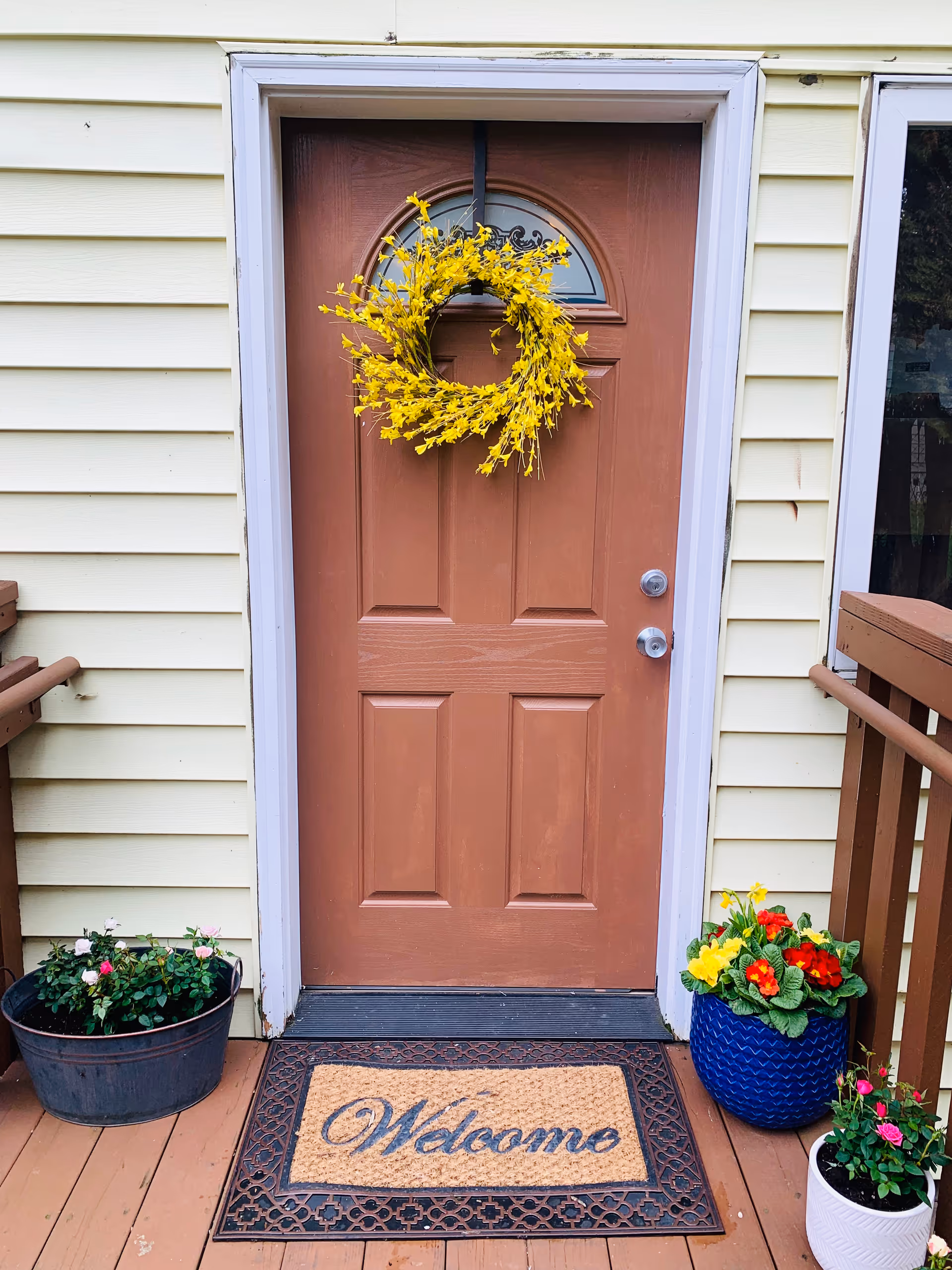 A brown front door with a yellow floral wreath hanging on it, surrounded by light yellow siding. There are three flower pots with colorful flowers on the wooden porch, and a welcome mat in front of the door.