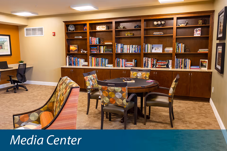 A cozy media center room with a round table surrounded by four chairs featuring colorful patterned upholstery. Behind the table is a large wooden bookshelf filled with books and decorative items. To the left, there is a small desk area with a black office chair and a framed picture on the wall. The room has beige walls and carpeted flooring.