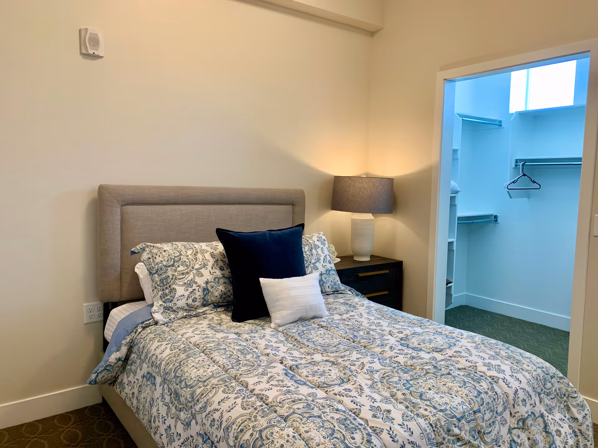 A bedroom with a bed featuring a beige upholstered headboard and patterned blue and white bedding. There are two pillows on the bed, one dark blue and one white. A nightstand with a lamp is next to the bed. An open doorway reveals a walk-in closet with shelves and hanging rods.