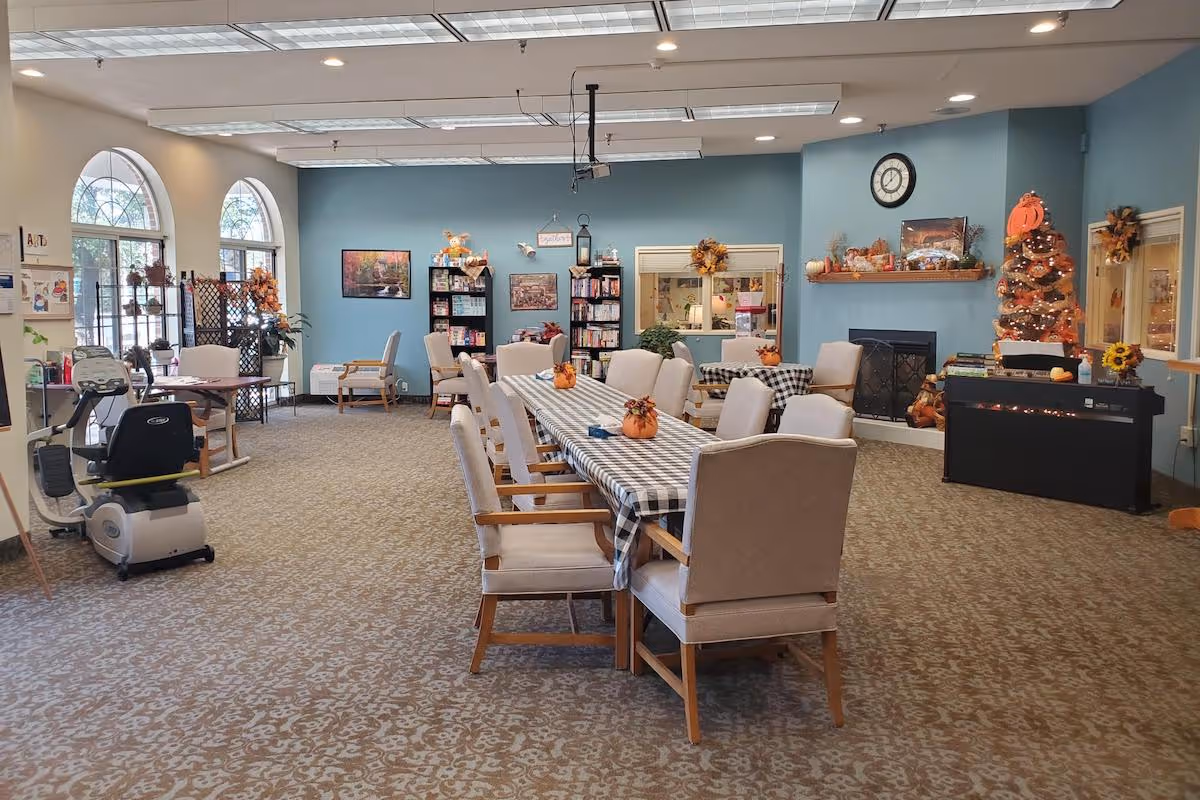 A spacious common area in a senior living facility with a long table covered in a checkered tablecloth and decorated with small pumpkin centerpieces. Comfortable chairs surround the table. The room features a fireplace with autumn decorations, a clock above it, bookshelves, and large arched windows letting in natural light. There is also an exercise machine and additional seating areas.