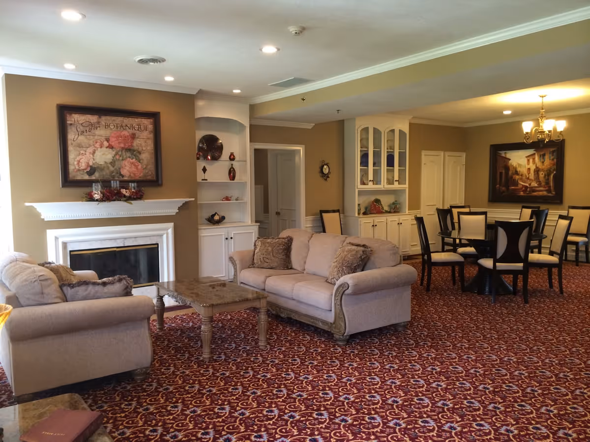A cozy living area in a senior living facility featuring beige sofas with decorative pillows, a marble-top coffee table, a fireplace with a floral painting above it, built-in shelves with decorative items, and a dining area with multiple tables and chairs under a chandelier. The room has warm beige walls and a patterned carpet.