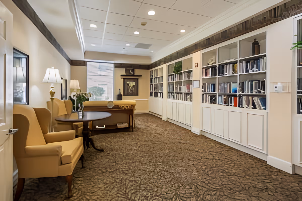 A cozy interior room with beige walls and carpeted floor, featuring a row of built-in white bookshelves filled with books and decorative items along the right wall. On the left side, there are two beige armchairs and a round wooden table with a small vase of flowers. The room is softly lit by table lamps and natural light coming through a window at the far end.