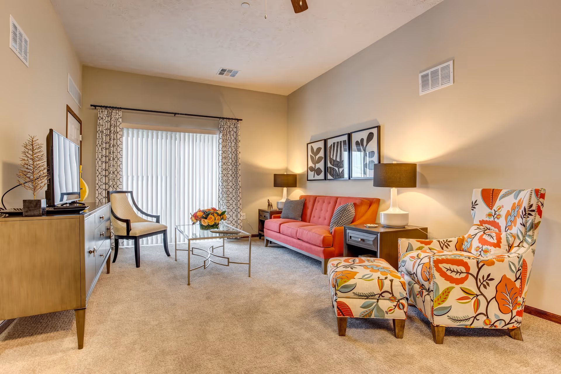 A cozy living room with a red sofa, two patterned armchairs with a matching ottoman, a glass coffee table with a flower arrangement, a TV on a wooden stand, and two table lamps on side tables. The room has beige walls, carpeted floor, and a large window with vertical blinds and patterned curtains.