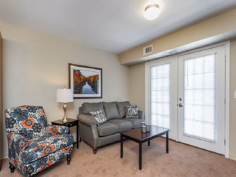 A cozy living room with a gray sofa adorned with two patterned pillows, a floral-patterned armchair, a dark wooden coffee table with decorative items, a side table with a lamp, and a framed picture of a river with autumn trees on the wall. There are double glass doors with blinds letting in natural light.