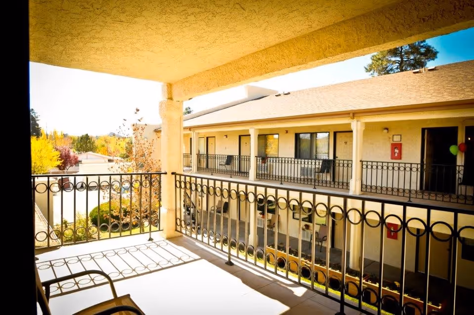 Sunlit second-floor balcony with decorative metal railing overlooking a courtyard and neighboring apartment doors at an assisted living facility.