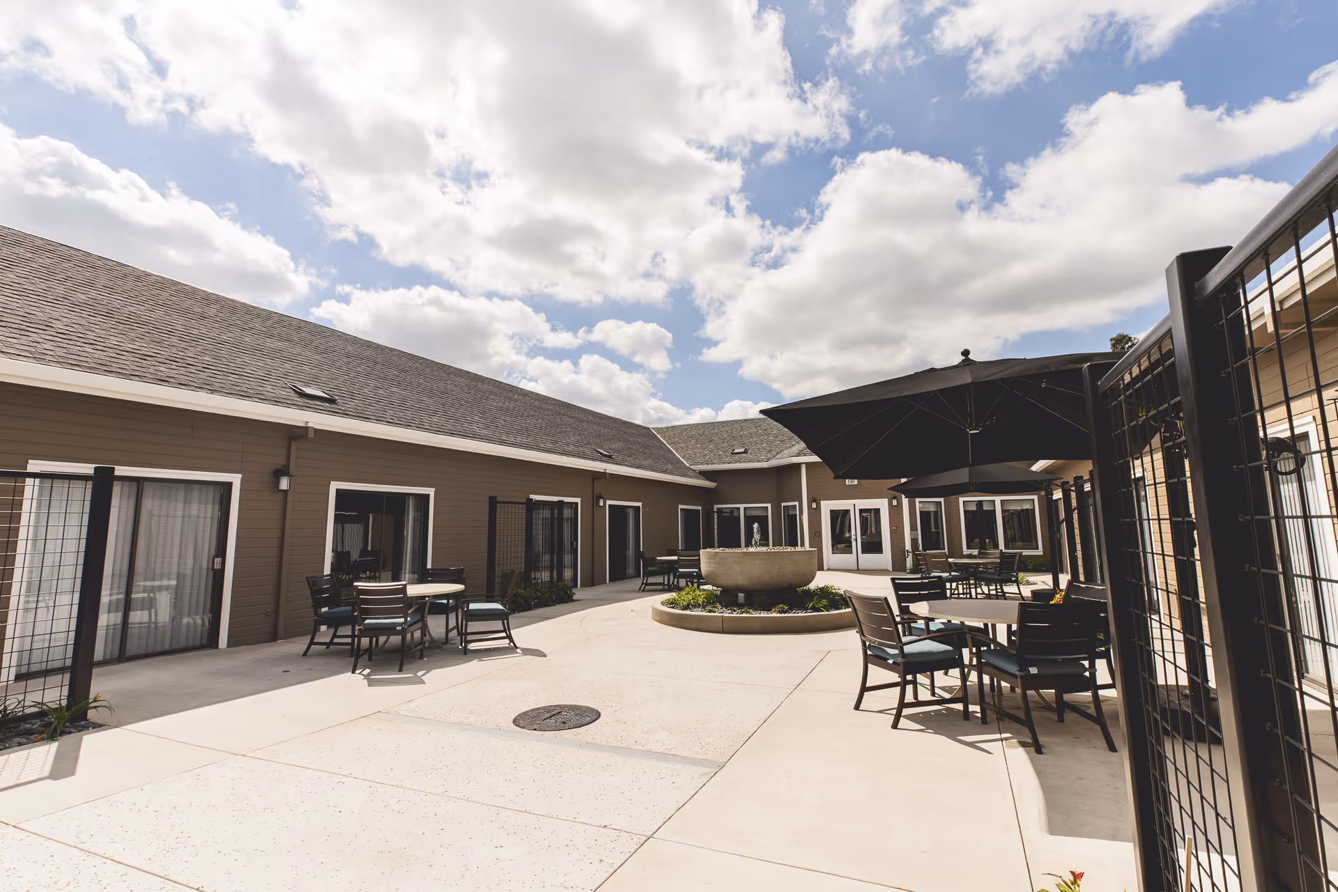 Outdoor courtyard area at Orangeburg Manor with several tables and chairs, some shaded by large black umbrellas. The courtyard is surrounded by a single-story building with multiple sliding glass doors. The sky is partly cloudy.