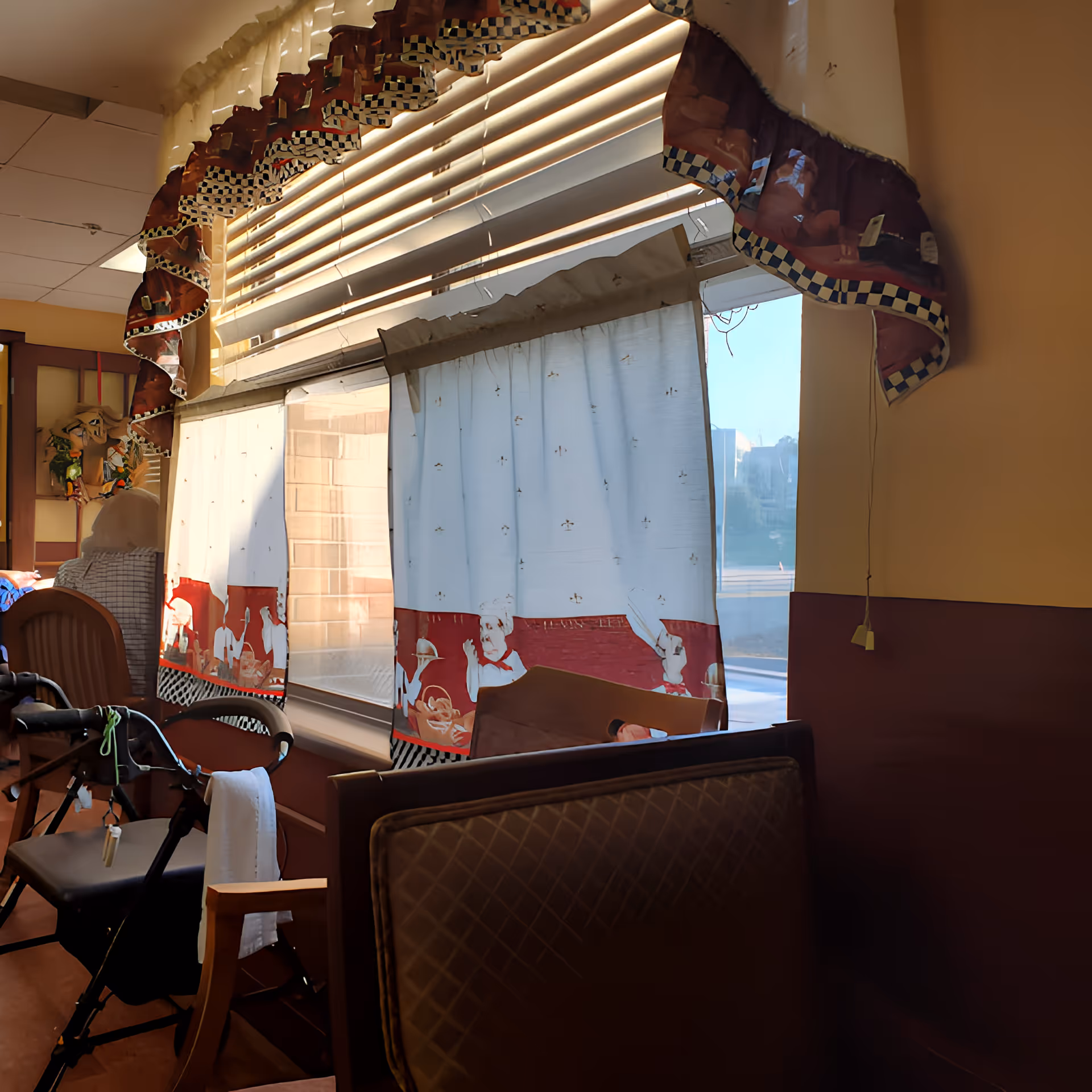 Dining-room interior with patterned valances and curtains over large windows, booth seating and a walker.