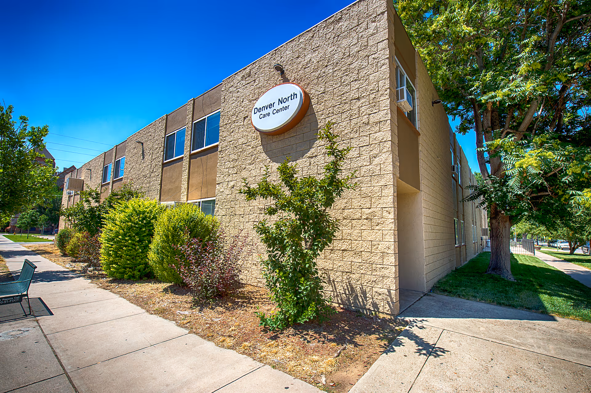 Exterior view of a two-story beige brick building with a sign that reads 'Denver North Care Center'. The building is surrounded by green bushes and trees, with a sidewalk and a bench visible in the foreground under a clear blue sky.