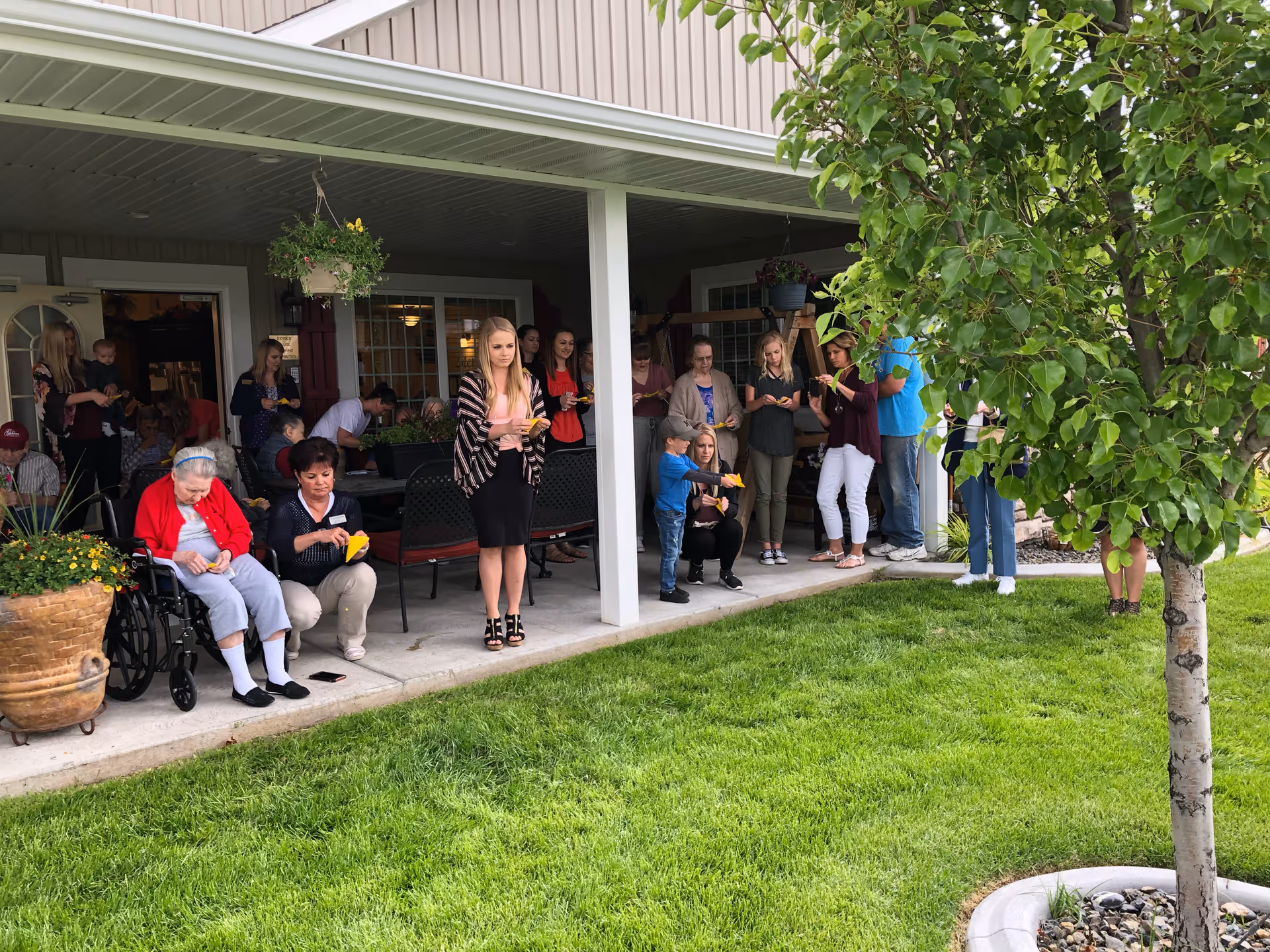 A group of people, including elderly individuals and caregivers, gathered outside under a covered patio area next to a green lawn and a tree. Some people are seated while others are standing, and many are holding yellow items, possibly food or napkins. The setting appears to be a social or community event at a senior living facility.