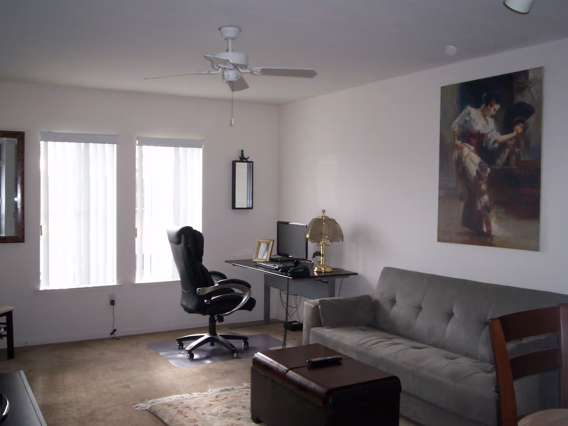A living room with a gray couch, a dark wooden chair, a small desk with a computer and a brass lamp, a black office chair on a plastic mat, two tall windows with vertical blinds, a ceiling fan, a framed painting of a woman holding a fan on the wall, and a small rug on the carpeted floor.