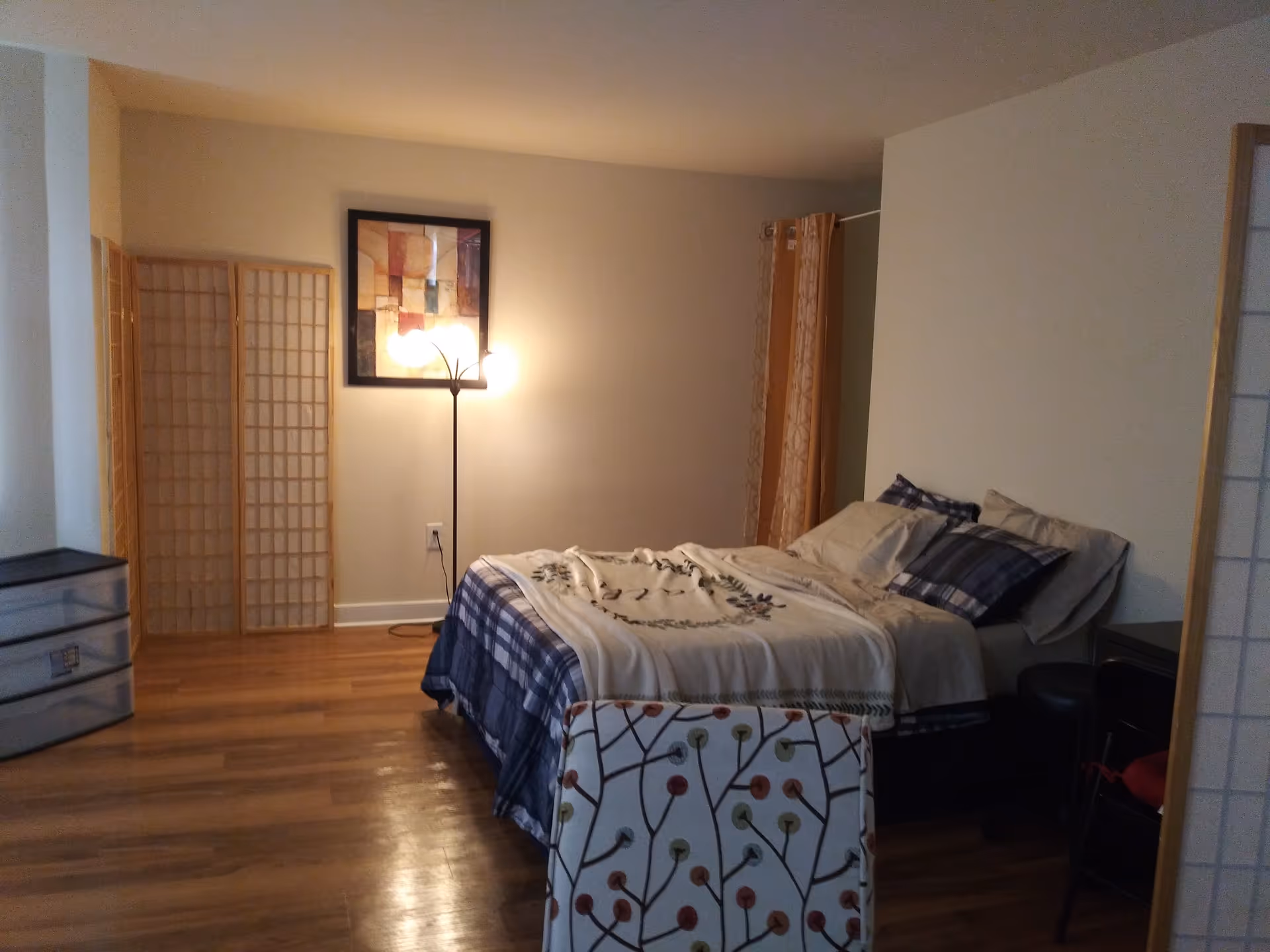 A furnished bedroom with a made bed, floor lamp, decorative room dividers, and hardwood floors.