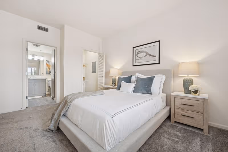 Bright, modern bedroom with a neatly made bed, two nightstands with lamps, and an open doorway leading to a bathroom.