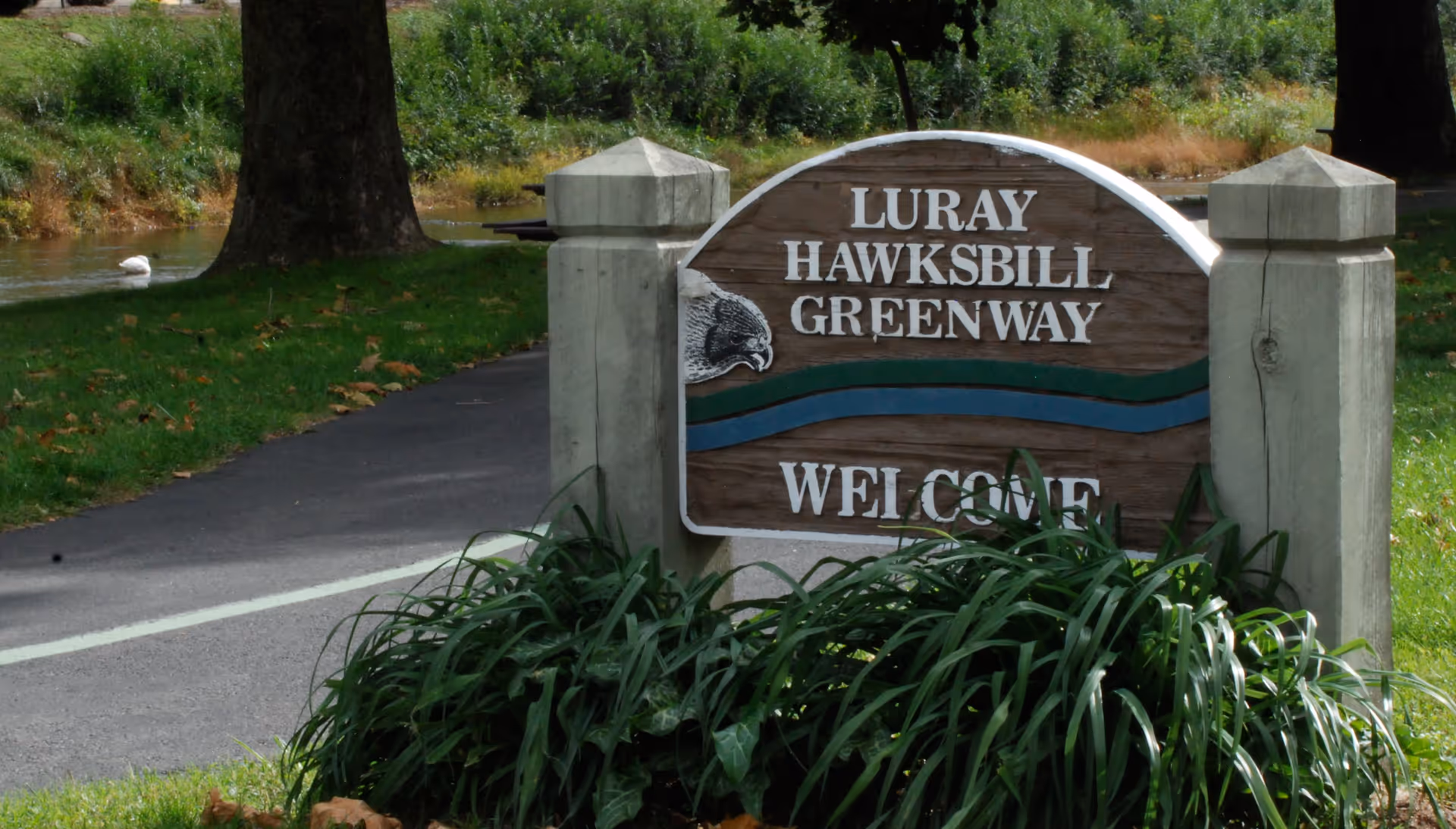 A wooden sign with white letters reading 'Luray Hawksbill Greenway Welcome' situated between two concrete posts, surrounded by green plants and grass, with a paved pathway and trees in the background.
