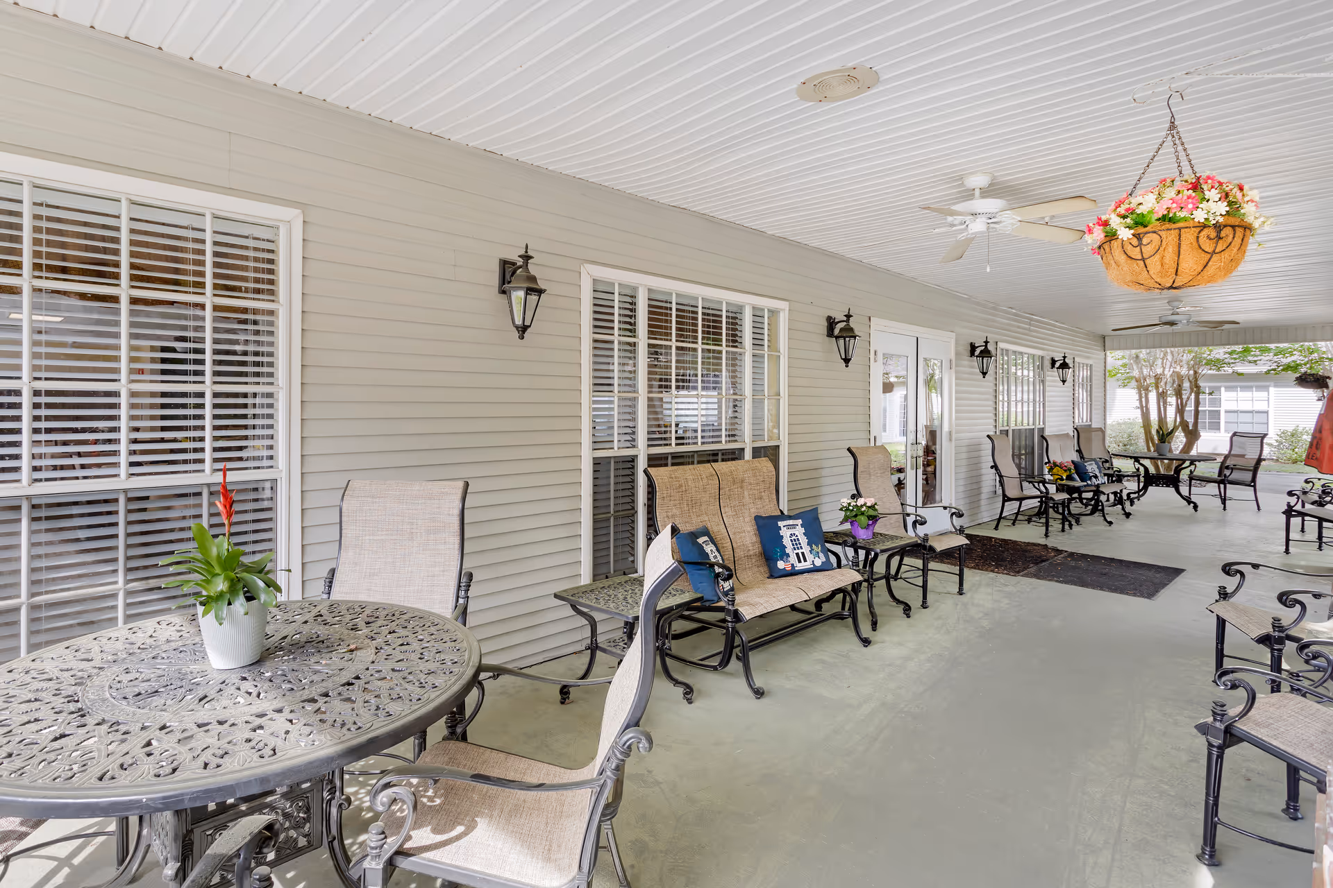 Covered outdoor patio area with metal tables and chairs, cushioned benches with decorative pillows, hanging flower baskets, ceiling fans, and wall-mounted lantern-style lights. Windows and doors of the building are visible along the wall.