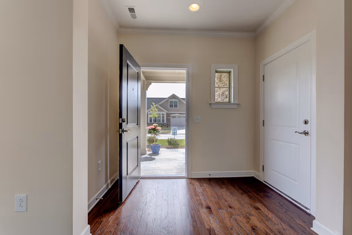 View of an entryway with an open black front door leading outside. The interior has beige walls, a small window to the right of the door, and dark hardwood flooring. Outside, a residential neighborhood with houses and greenery is visible.