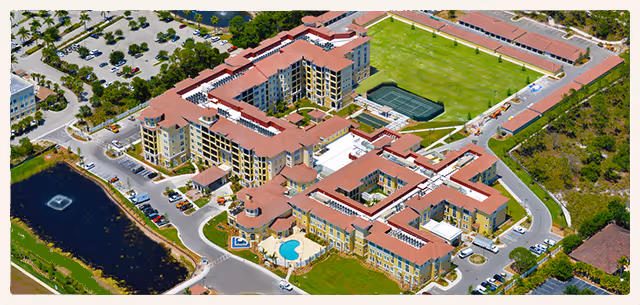 Aerial view of The Terraces at Bonita Springs senior living facility showing multiple connected buildings with red roofs, a swimming pool, tennis courts, parking areas, and surrounding greenery.