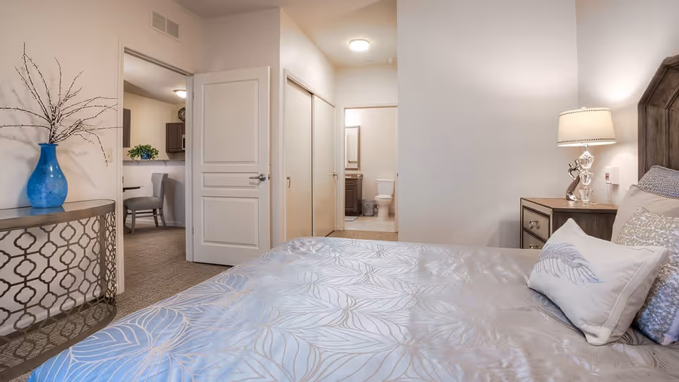 A bedroom with a bed covered in a light blue patterned bedspread and decorative pillows. A wooden nightstand with a lamp is beside the bed. To the left, there is a decorative table with a blue vase holding branches. The room opens to a hallway leading to a bathroom with a visible toilet and sink. A dining area with a chair and kitchen cabinets is partially visible through an open door.