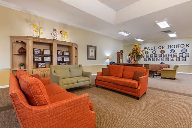 A senior living facility common area with orange and green upholstered sofas arranged around a carpeted floor. A wooden cabinet with framed photos and decorative items is against the wall. In the background, a 'Wall of Honor' displays photos and plaques. The room is well-lit with ceiling lights and a wall lamp.