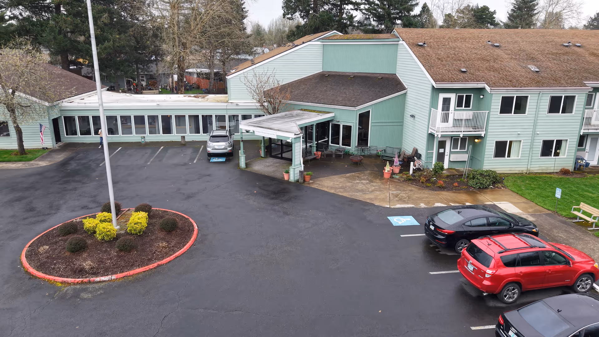 Exterior view of a two-story green retirement living facility with a covered entrance, several parked cars, a small landscaped island with bushes and a flagpole, and a bench near the sidewalk.