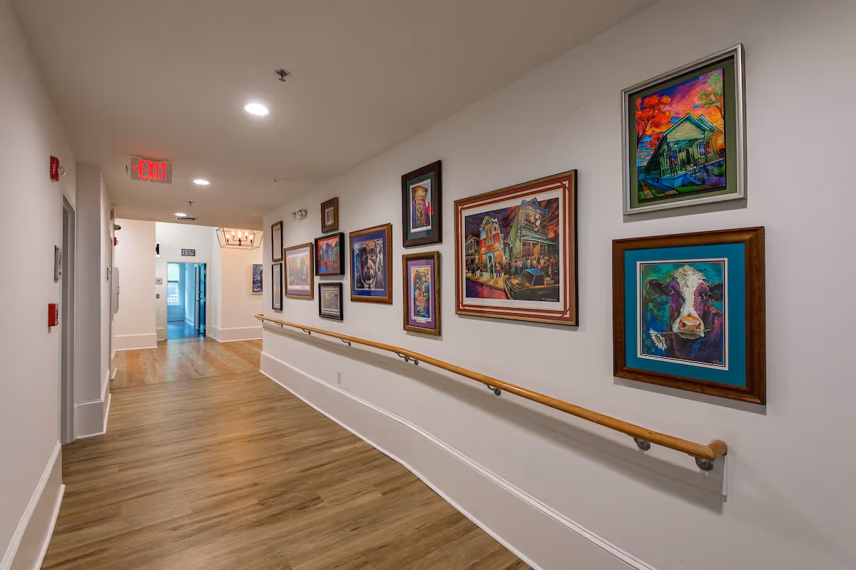 A well-lit hallway with wooden flooring and white walls adorned with colorful framed paintings. A wooden handrail runs along the right side of the hallway. At the end of the hallway, there is an exit sign and a doorway leading to another room.