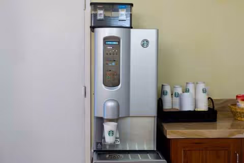 Corner interior beverage station with a coffee machine, cups, and a wooden counter.