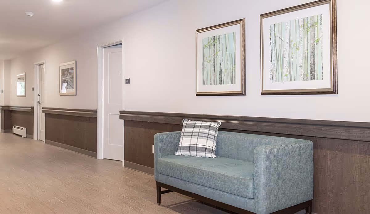 A hallway in a senior living facility with light-colored walls and wood paneling along the lower half. There are several white doors along the hallway, framed nature-themed artwork on the walls, and a blue upholstered bench with a plaid cushion placed against the wall.