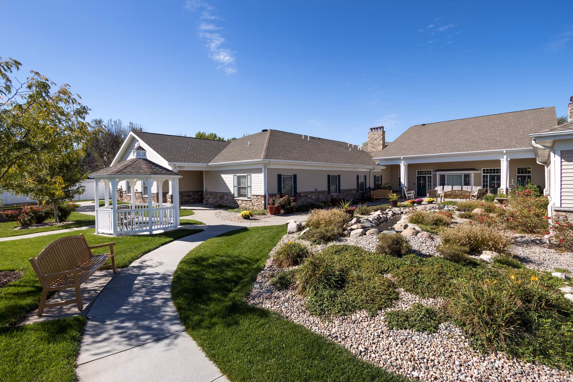 Outdoor view of a senior living facility with a white gazebo, benches, a landscaped garden with rocks and shrubs, and a building with a covered porch under a clear blue sky.