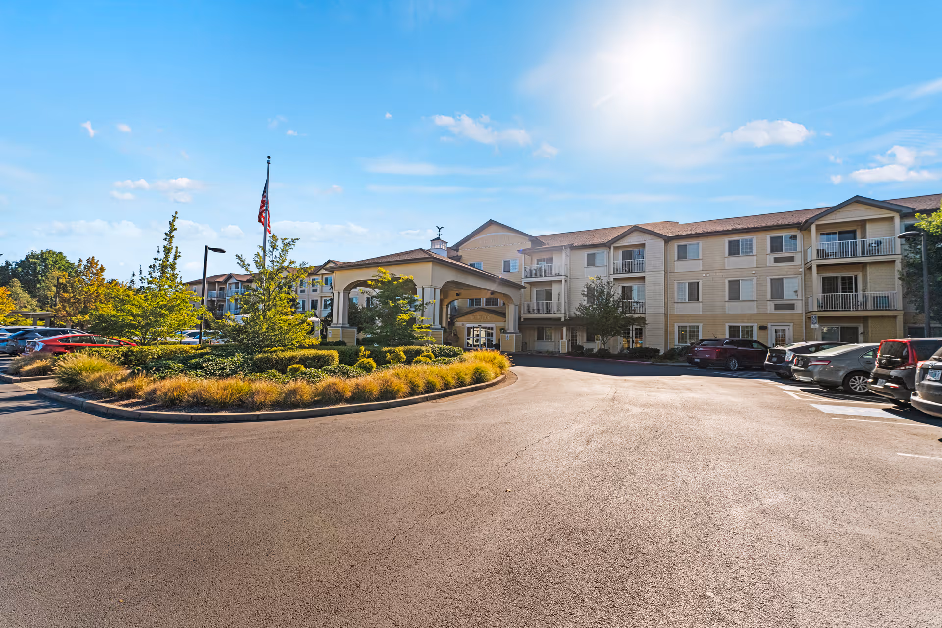 Exterior view of Volante Senior Living of Eugene, showing a three-story building with balconies, a covered entrance, landscaped greenery, parked cars, and a clear blue sky with the sun shining.