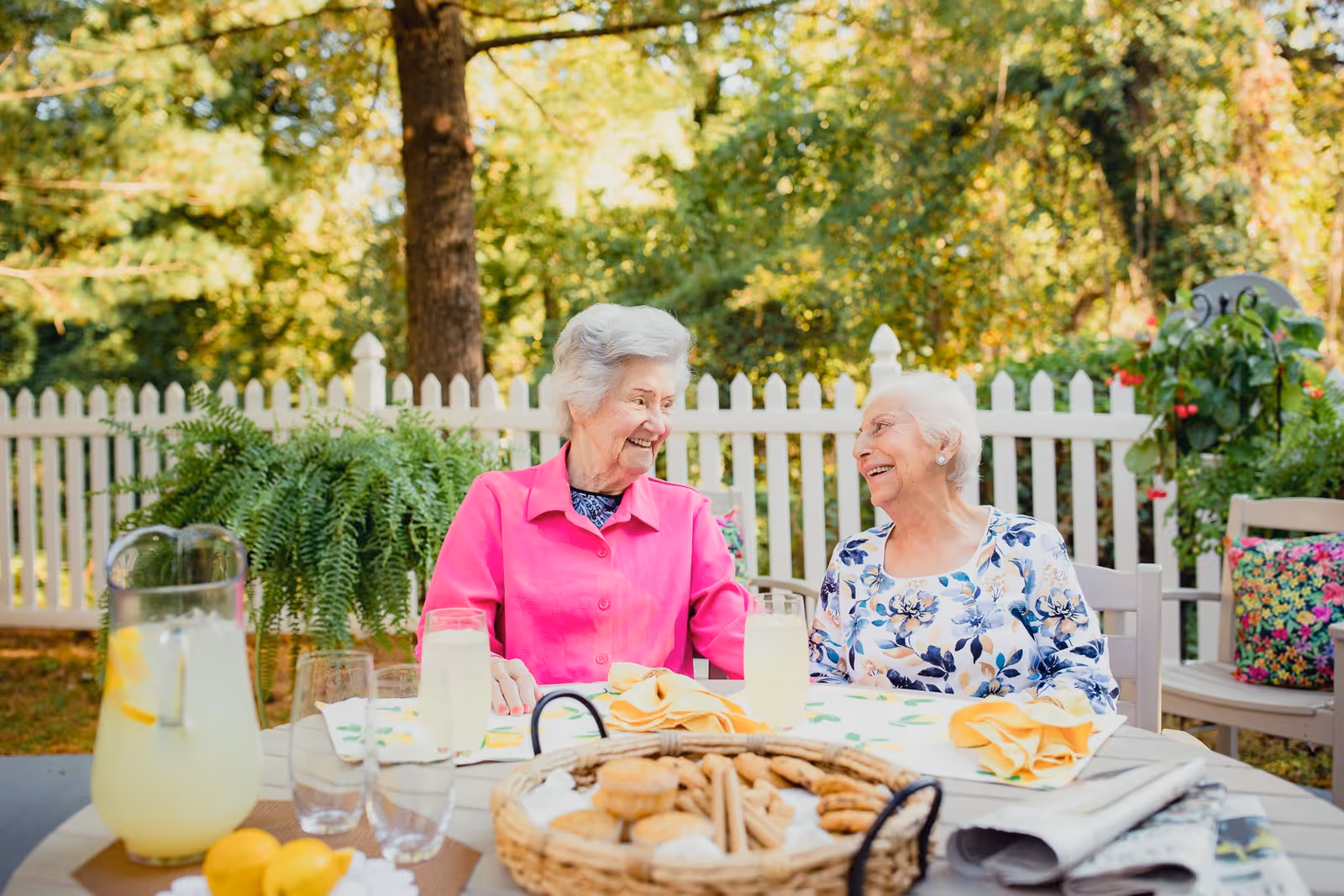 Two elderly women sitting at an outdoor table with lemonade and a basket of cookies, smiling and enjoying a sunny day in a garden with a white picket fence and green trees in the background.