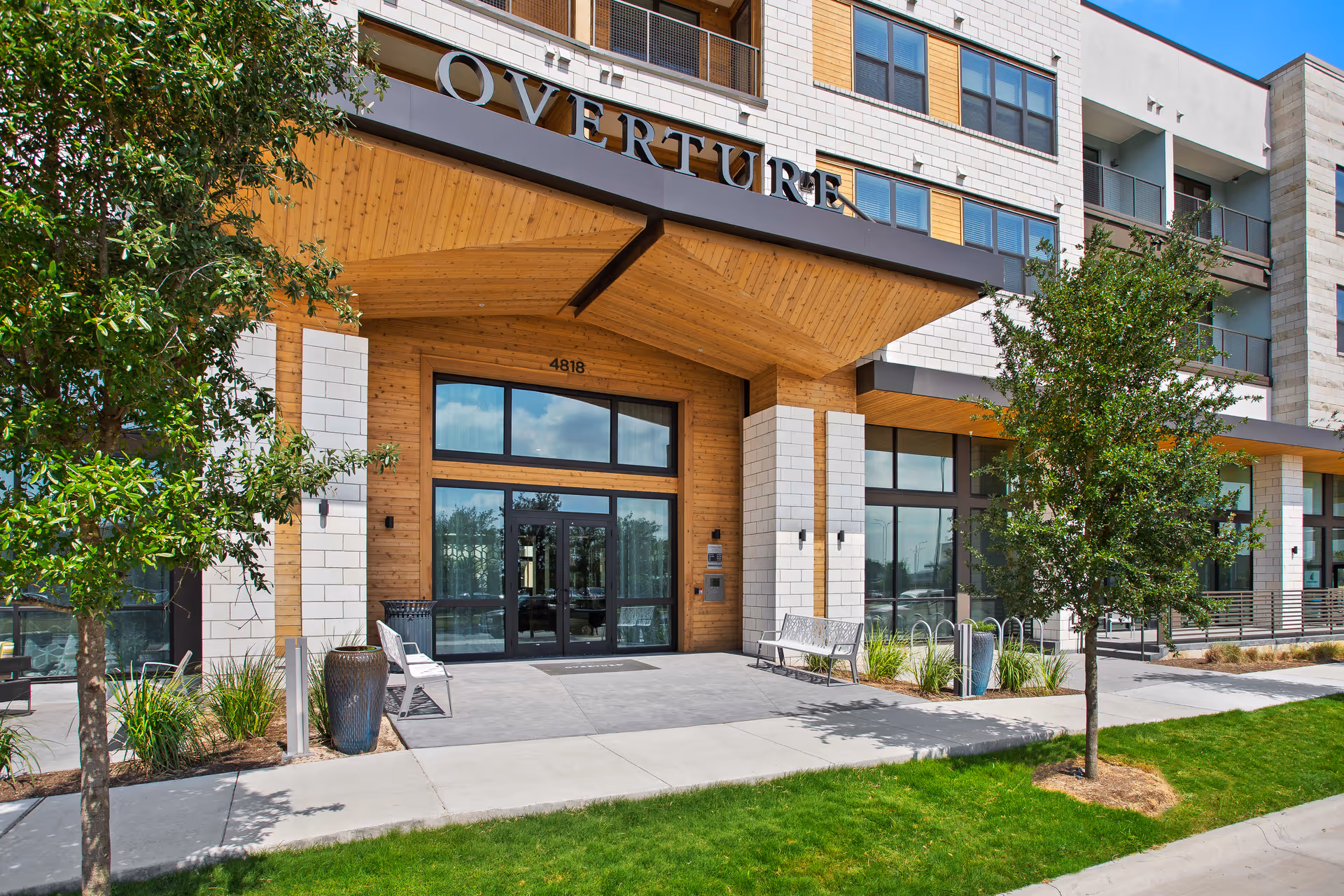 Entrance of Overture Mueller senior living facility featuring a modern facade with wood and white brick accents, large glass doors, benches on either side, small trees, and a well-maintained lawn.