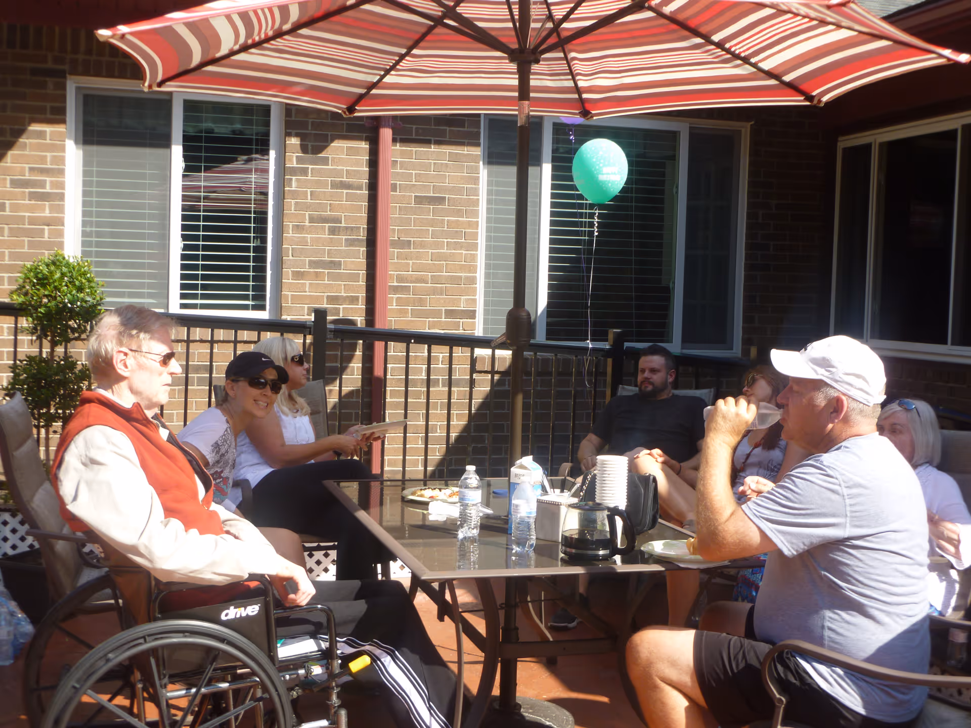 Several people, including an older man in a wheelchair, seated around a patio table under a striped umbrella in an outdoor patio area.
