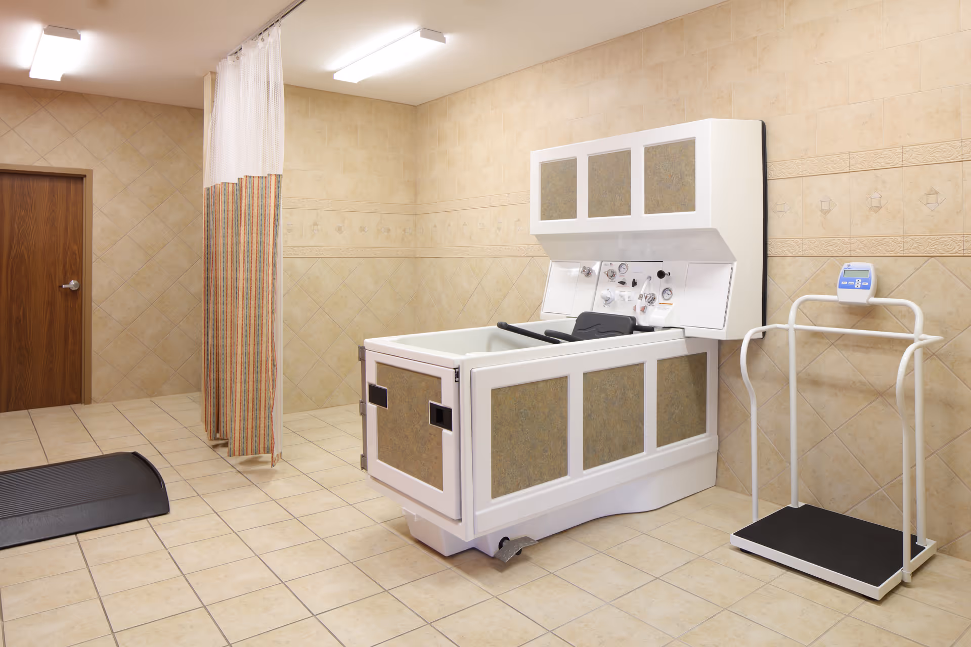 A tiled room with a medical bathing tub equipped with controls and a chair, a weighing scale with handrails, a privacy curtain, and a wooden door. The walls and floor are covered with beige tiles.