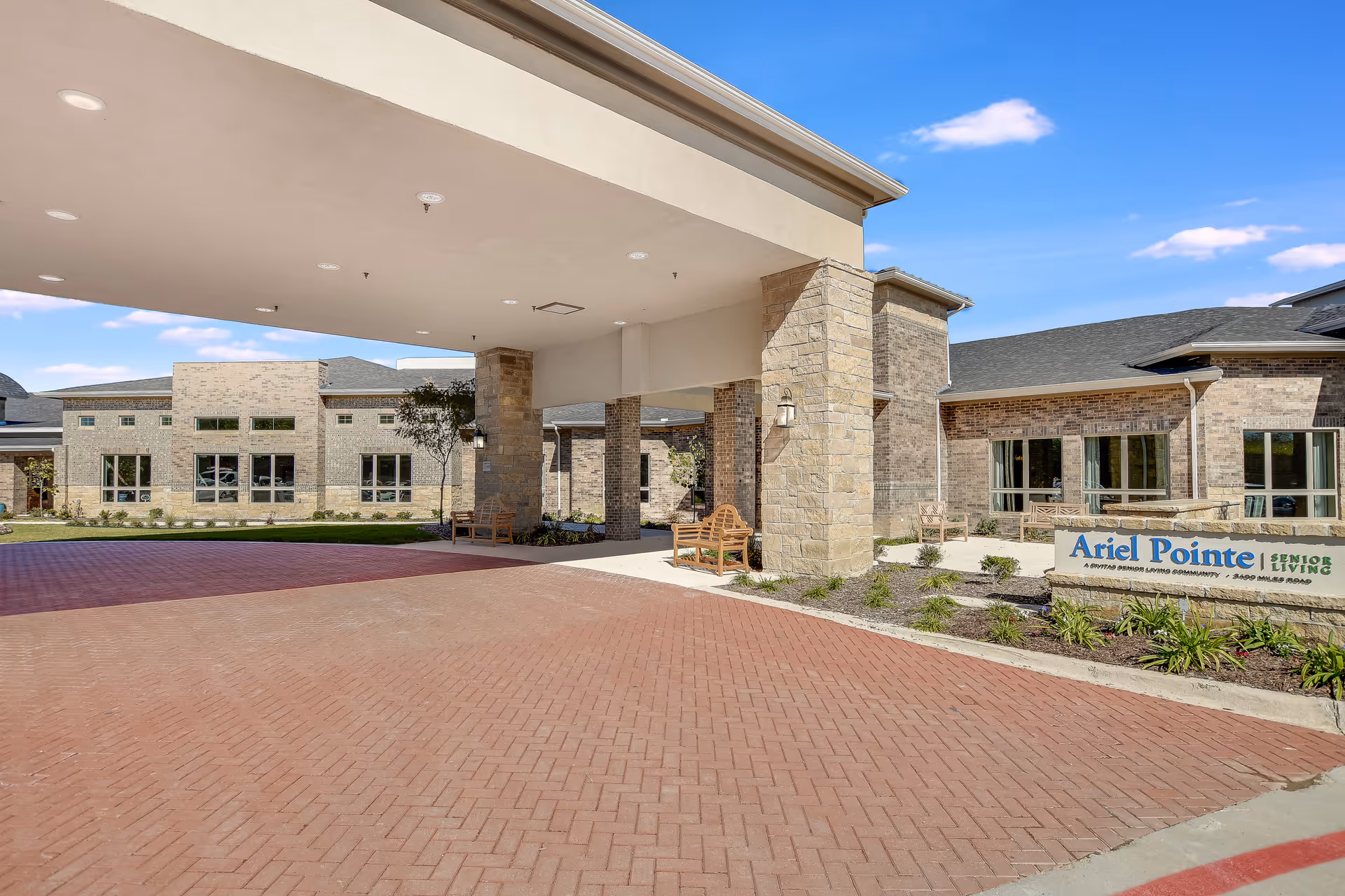 Exterior view of Ariel Pointe of Sachse Senior Living facility showing a covered entrance with stone pillars, benches, and a brick driveway under a clear blue sky.