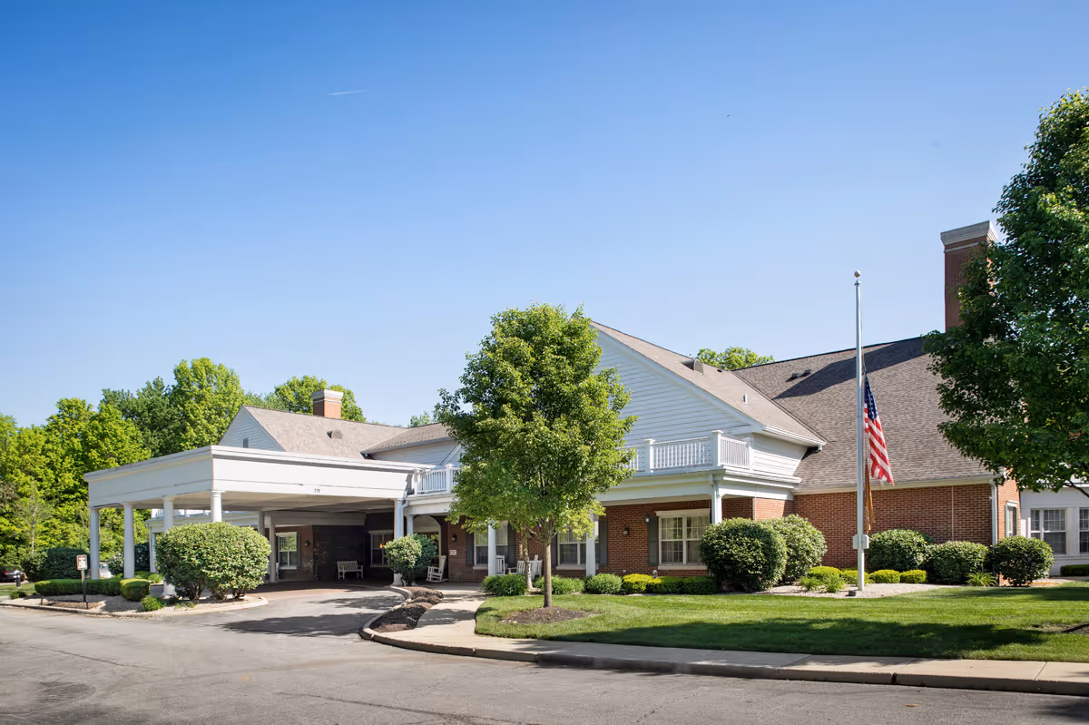 Exterior view of a senior living facility building with a covered entrance, well-maintained landscaping including bushes and trees, and an American flag on a flagpole under a clear blue sky.