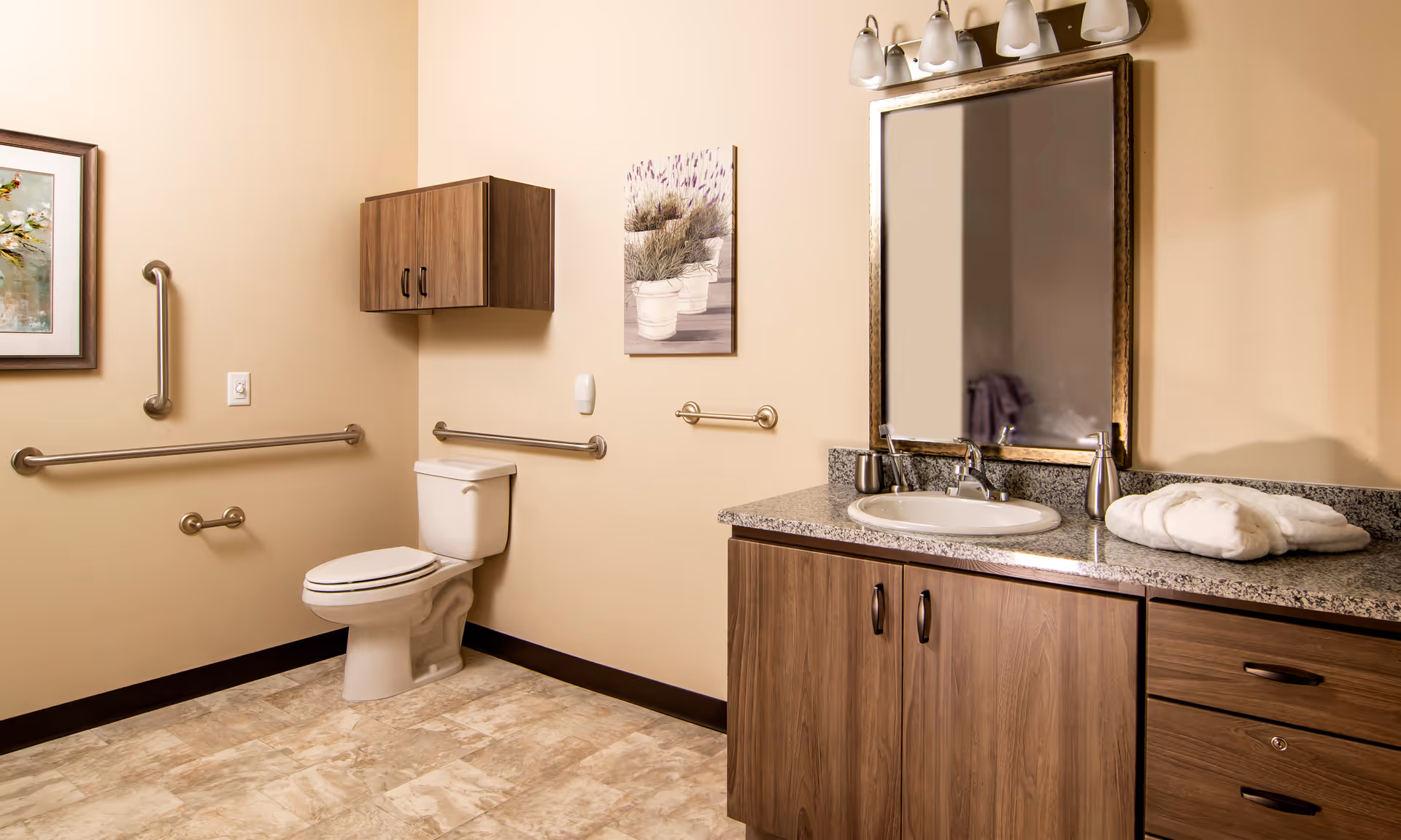 A clean and accessible bathroom with beige walls and tiled floor. The bathroom features a white toilet with grab bars on the walls around it, a wooden cabinet mounted above the toilet, and a wooden vanity with a granite countertop, sink, and a large mirror above it. There are folded white towels on the countertop and two framed pictures on the walls.