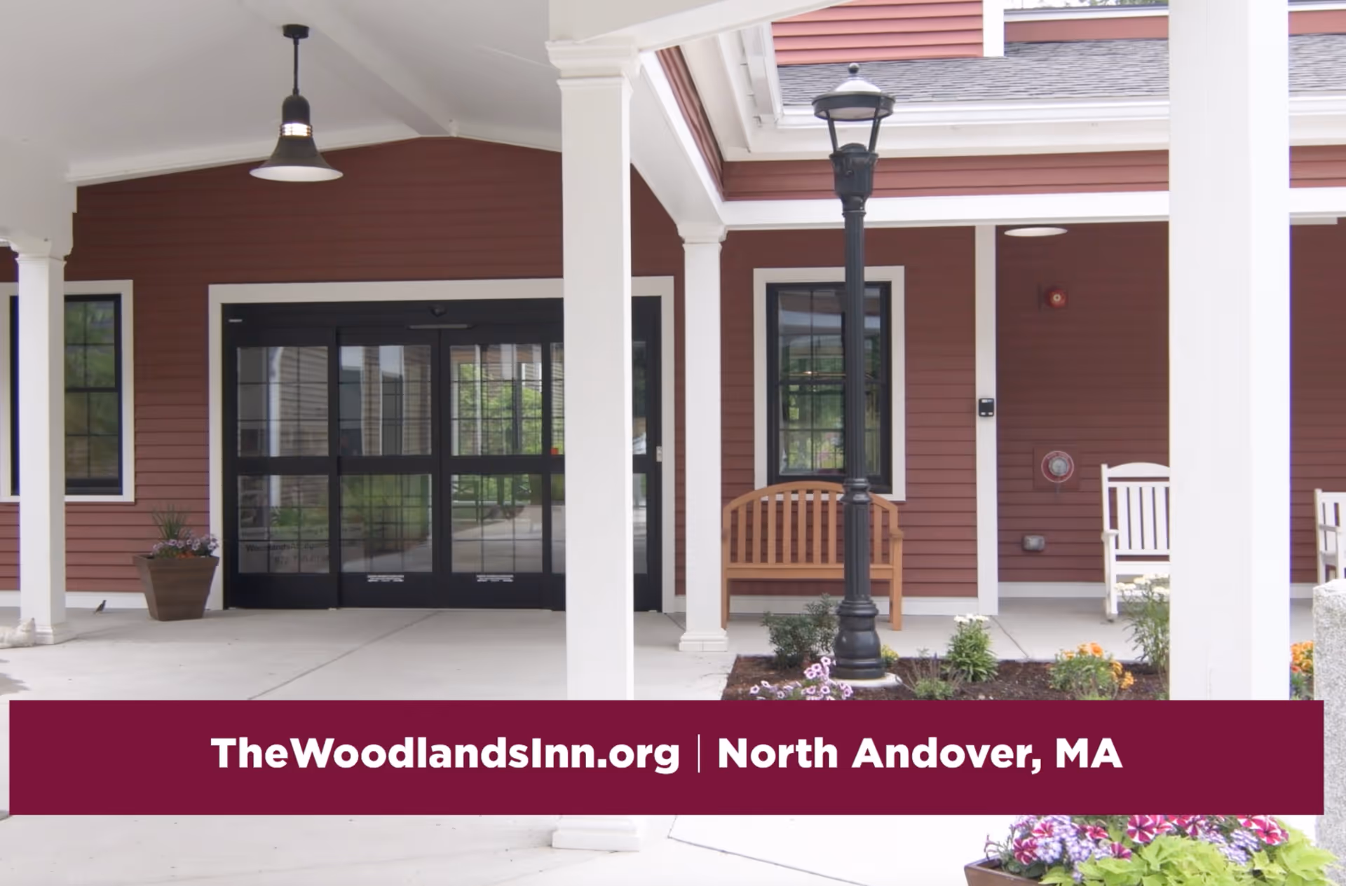 Covered entrance area of a building with red siding and white trim, featuring a black automatic sliding door, a wooden bench, a black street lamp, potted plants, and a hanging light fixture.