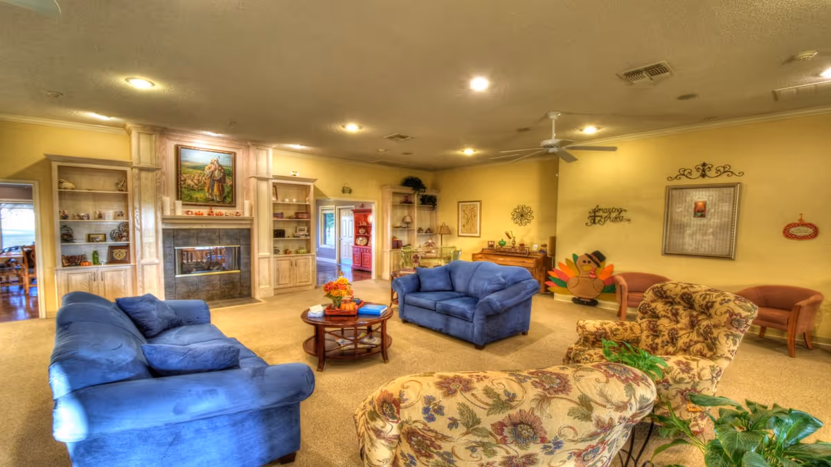 A cozy living room in a senior living facility with two blue sofas, two floral armchairs, a round wooden coffee table with flowers and books, a fireplace with built-in shelves on either side, and various decorative items on the walls and shelves. The room has warm yellow walls and carpeted flooring.