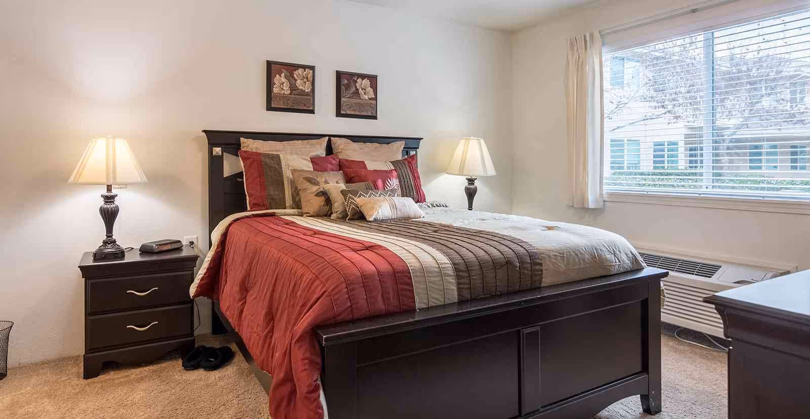 A neatly made bed with a dark wooden frame and a red, beige, and brown striped comforter in a bright bedroom. There are multiple pillows arranged on the bed. On either side of the bed are matching dark wooden nightstands with lamps. A large window with white blinds and curtains lets in natural light, and there are two framed floral artworks on the wall above the bed.