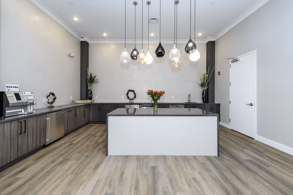 Modern kitchen area with a large central island topped with a vase of red flowers, surrounded by dark wood cabinets and countertops. Multiple pendant lights with different shapes and colors hang from the ceiling. The floor is light wood, and there is a white door on the right side of the room.