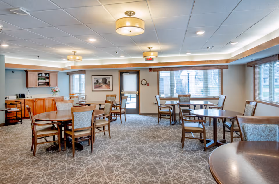A bright and spacious dining room with several round wooden tables and cushioned chairs arranged neatly. The room has large windows with blinds allowing natural light to enter, a carpeted floor with a patterned design, and ceiling lights with drum-shaped shades. There is a wooden cabinet with shelves and a microwave on the left side, and a door at the back leading outside.