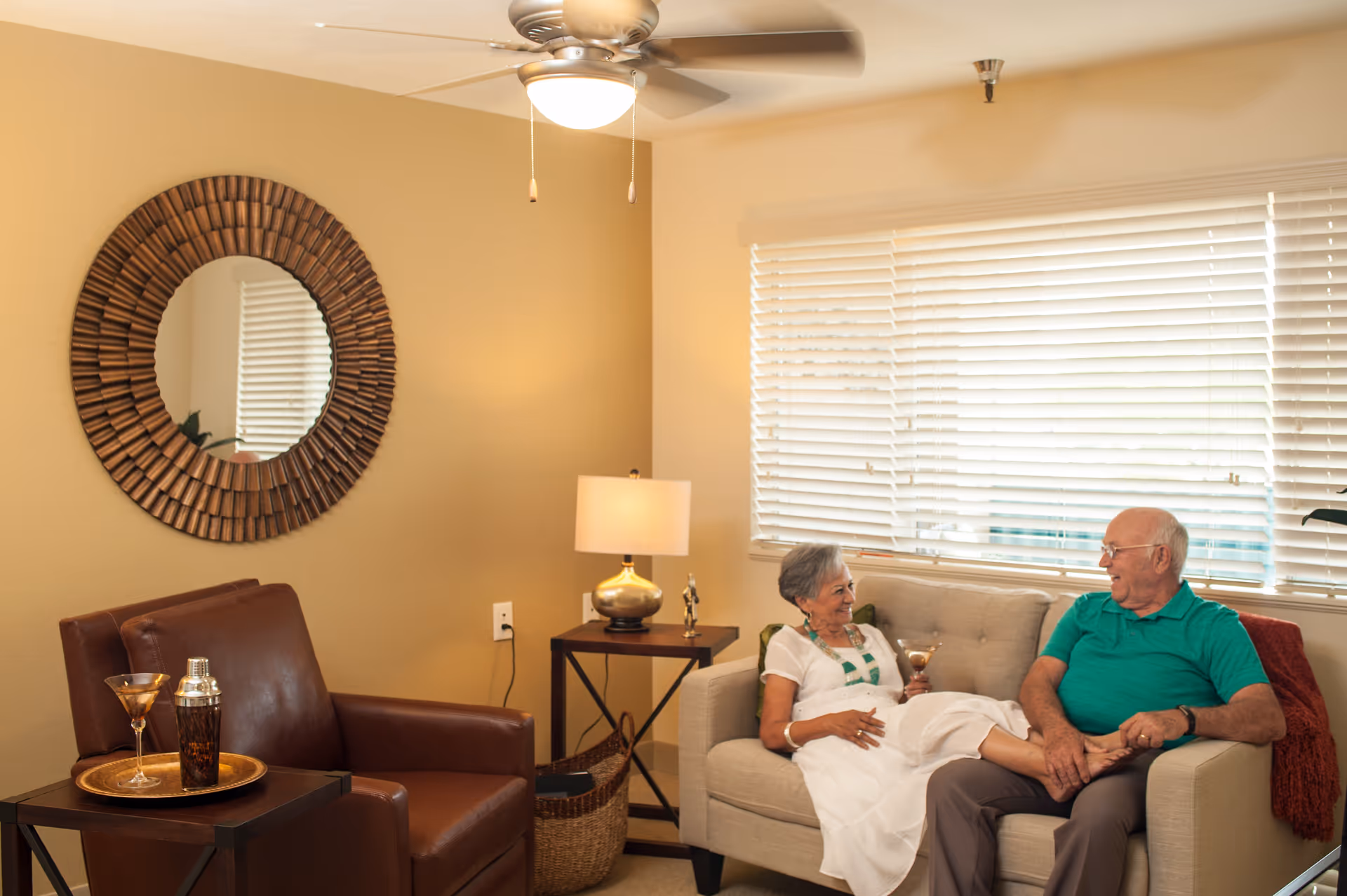 An elderly couple sitting and relaxing in a cozy living room. The woman is seated on a beige couch holding a drink, while the man sits beside her, holding her foot. The room features a brown leather armchair with a tray holding a cocktail shaker and a glass, a side table with a lamp, a round decorative mirror on the wall, and a ceiling fan above. Light streams in through a window with white blinds.
