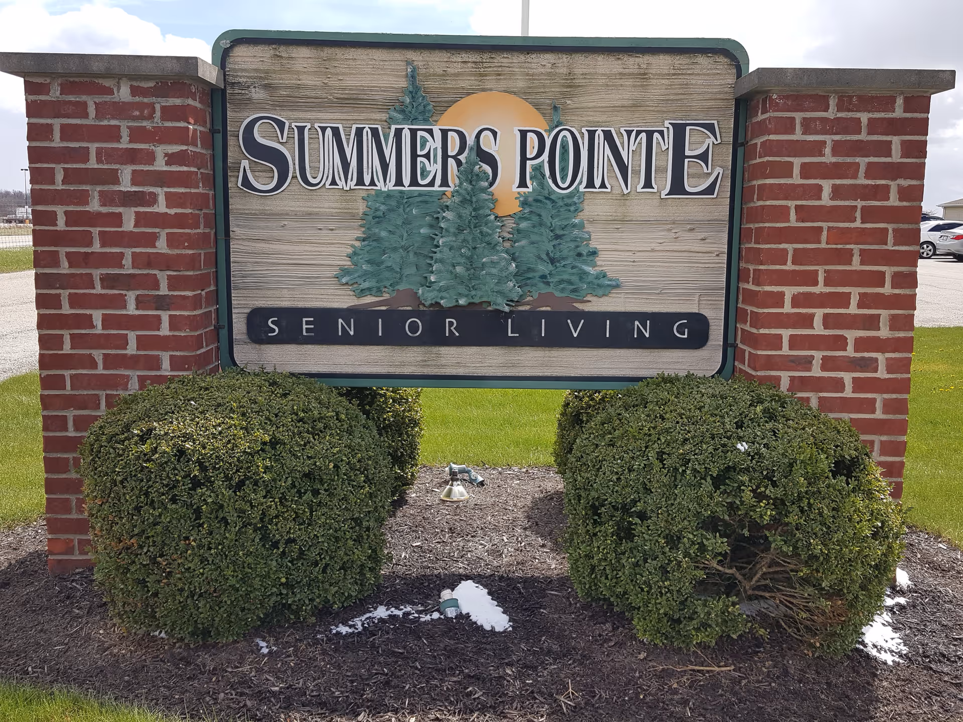 A wooden sign mounted between two red brick pillars with green bushes at the base. The sign reads 'Summers Pointe Senior Living' with an illustration of three evergreen trees and a sun in the background. The sign is outdoors on a grassy area with a parking lot and cars visible in the background.