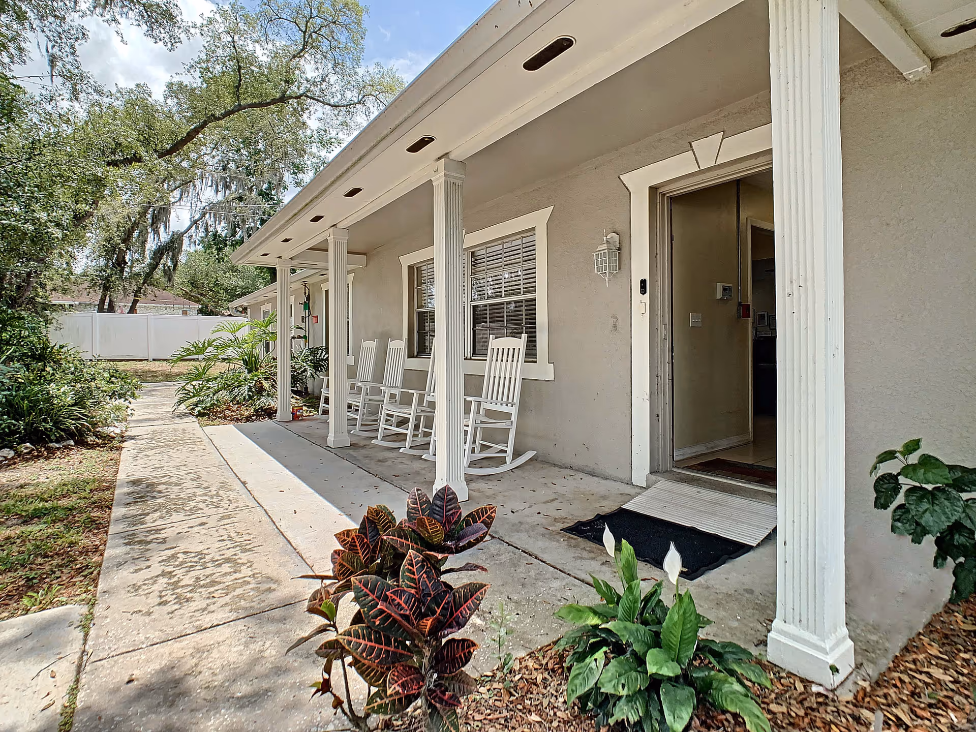 Outdoor view of a porch area at Park View Estate of Brandon ALF featuring four white rocking chairs lined up against the building wall. The porch is supported by white columns and has a concrete walkway leading up to it. There are various green plants and shrubs around the porch, and a white fence is visible in the background under a partly cloudy sky.