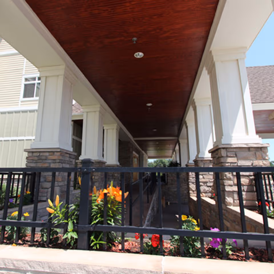 Covered outdoor walkway with white columns and stone bases, black metal railing, and colorful flowers planted along the sides under a wooden ceiling.